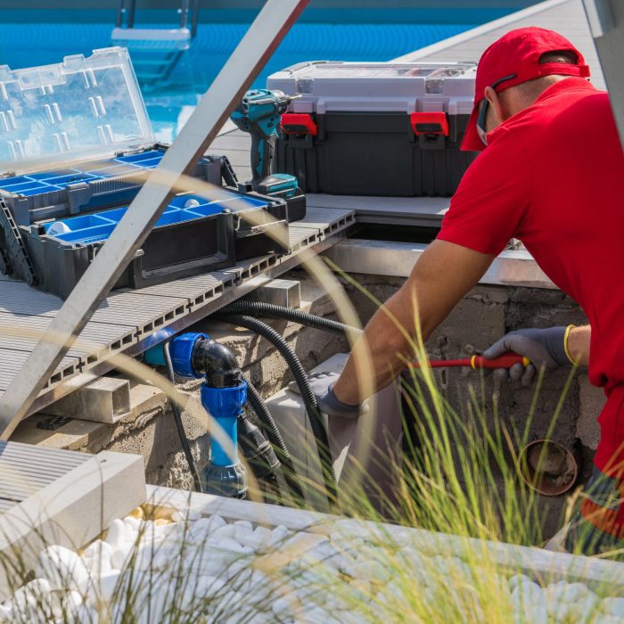 Closeup of Caucasian Professional Worker in Red Uniform Fixing Outdoor Swimming Pool Heating System Using Different Tools. Side View.